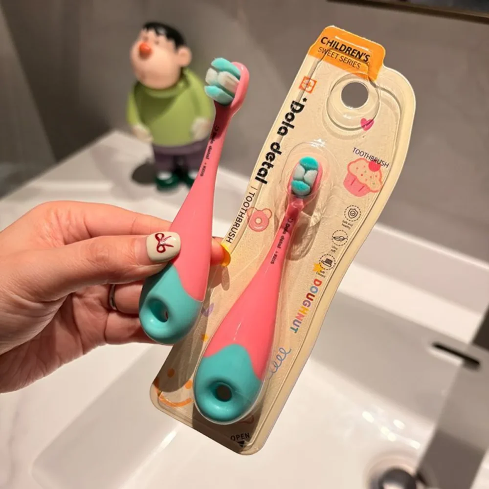Hand holding pink-mint Grin Teeth KT 3 donut toddler toothbrush in blister pack by sink; BPA-free body, ultra-soft bristles
