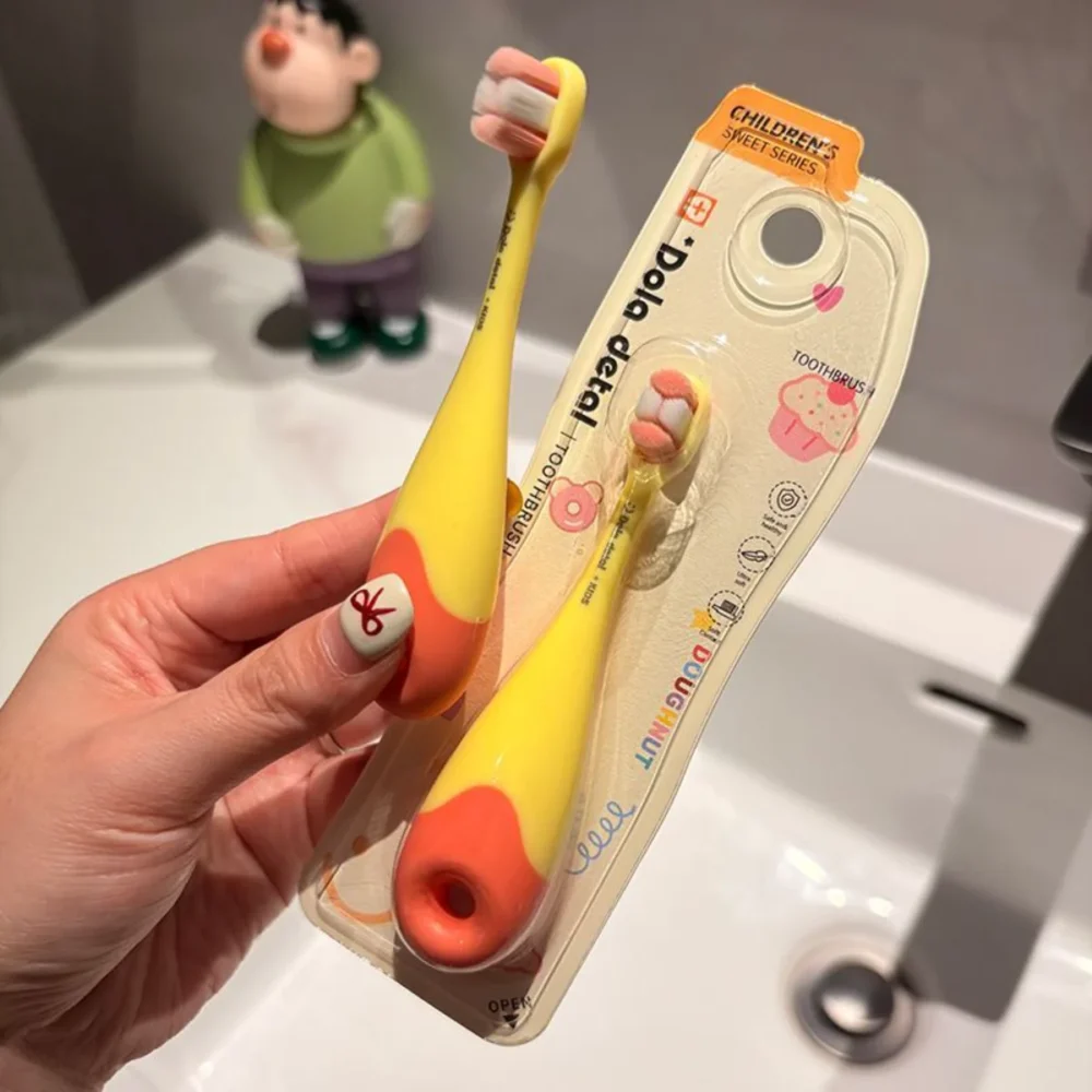 Hand holding Grin Teeth KT 3 donut toddler toothbrush—yellow donut handle, ultra-soft bristles, blister pack beside sink