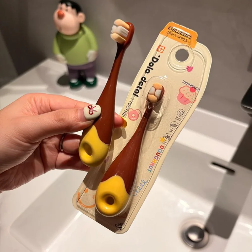 Hand holding brown-yellow Grin Teeth KT 3 donut toddler toothbrush in blister pack beside sink; BPA-free, ultra-soft bristles