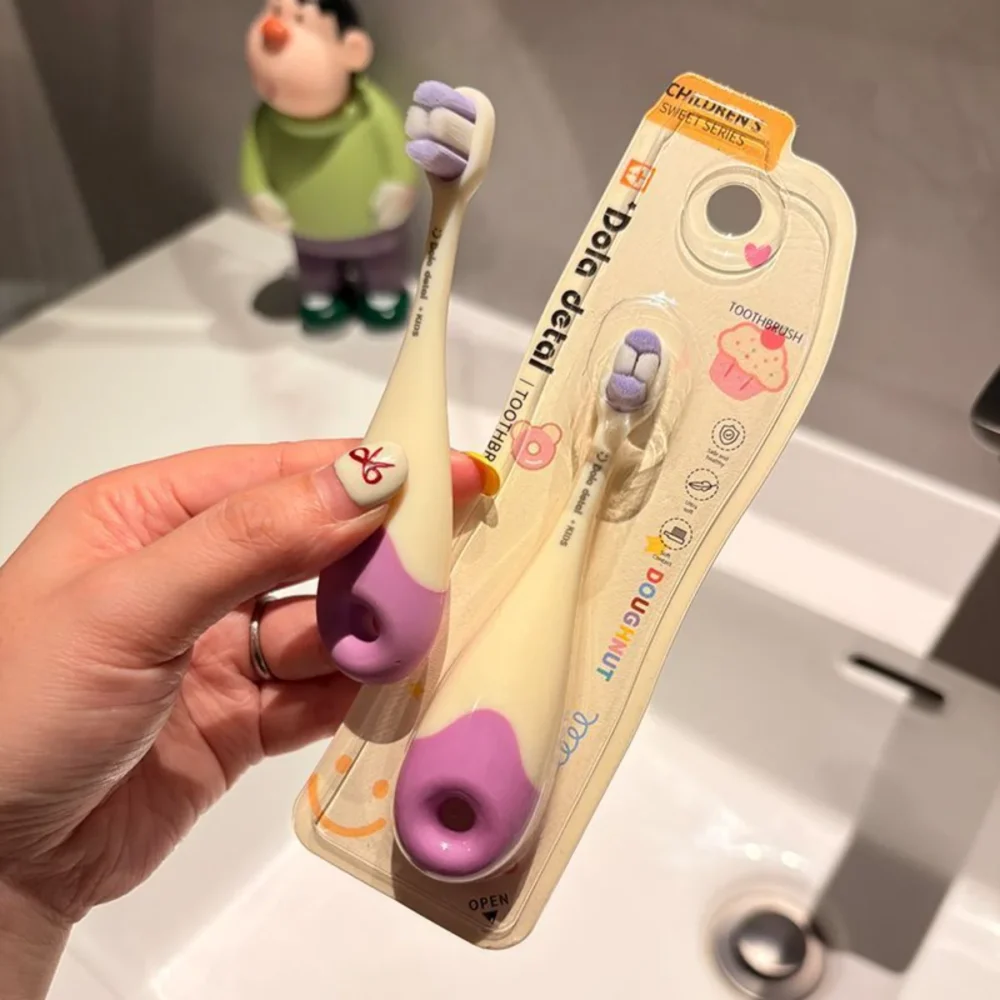 Hand holding ivory-lavender KT 3 donut toddler toothbrush in blister pack; BPA-free, extra-soft bristles, donut loop handle