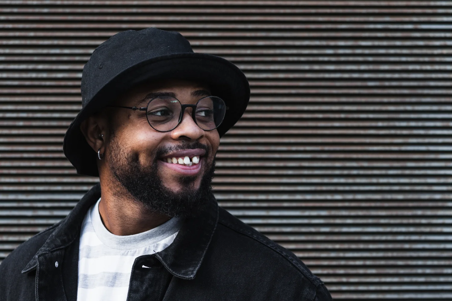 Grin Teeth About Us – Real People Confident Smile Grin Teeth About Us image: smiling man in glasses and hat with a natural gap-tooth smile against a striped wall