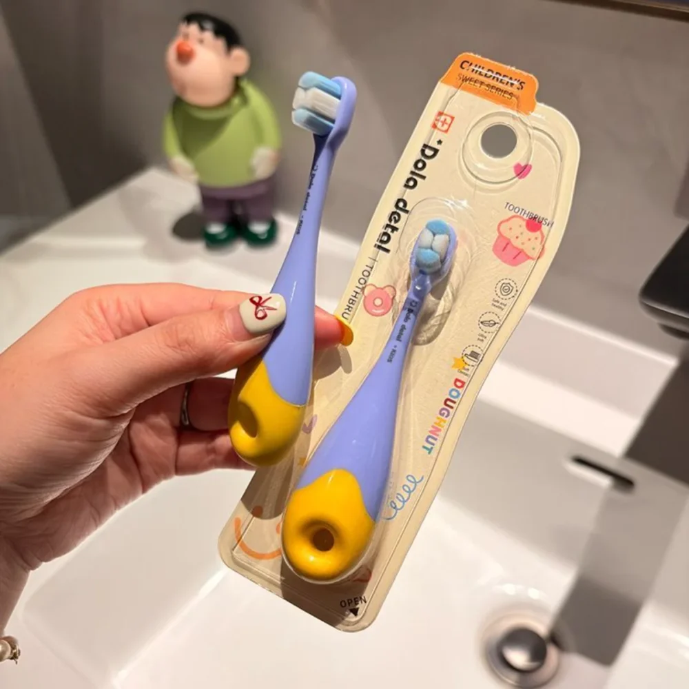 Hand holding blue-yellow Grin Teeth KT 3 donut toddler toothbrush in pack by sink; food-grade body, feather-soft bristles